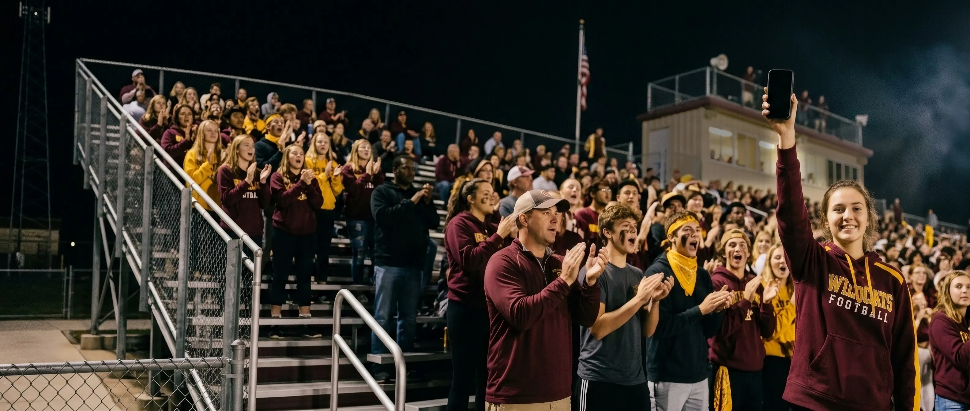 A fan in the home stands at a night high-school football game smiles with her phone held high among cheering, face-painted classmates. Go Wildcats! sweeps along the arc of her wave in white letters outlined in yellow.