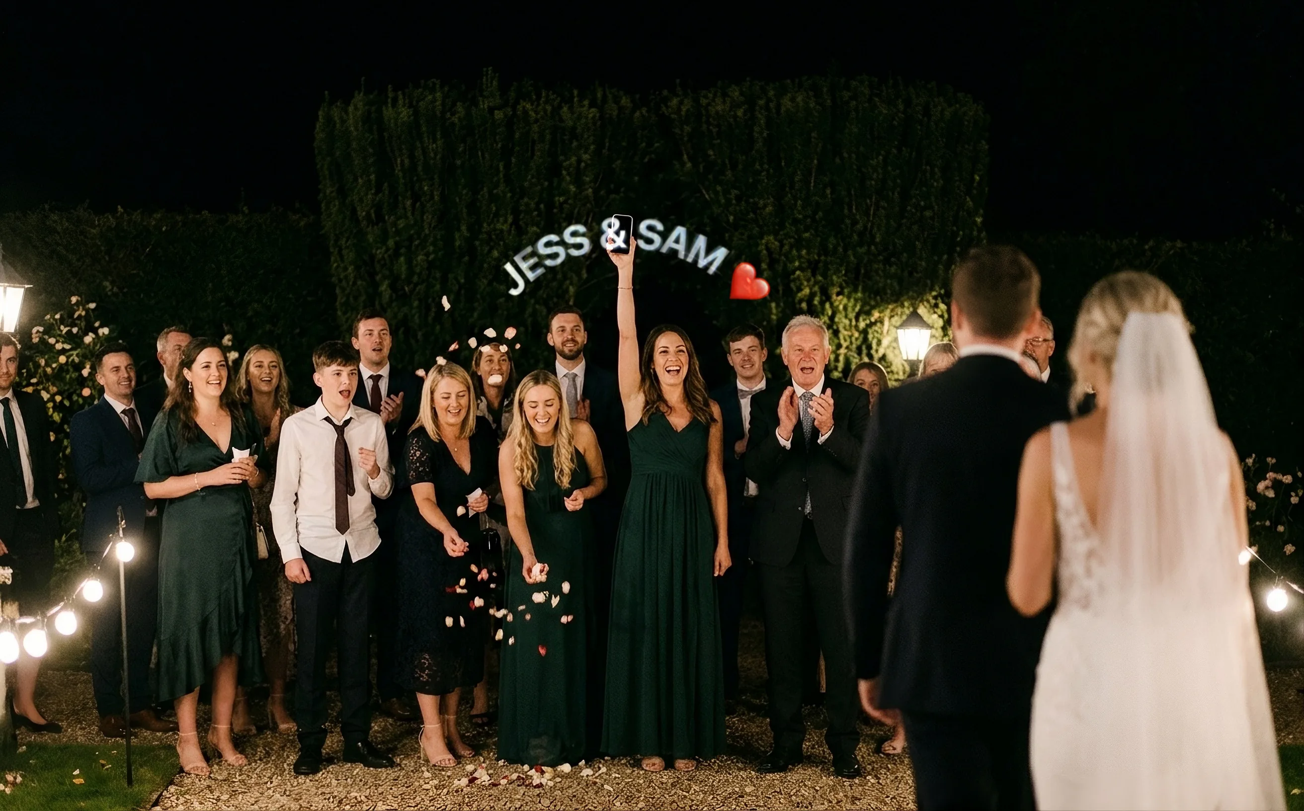 Wedding guests lining a garden path at night cheer as the bride and groom walk toward them. A woman in a green dress holds her phone high, and JESS & SAM, with a red heart, arcs above her arm in glowing white letters.